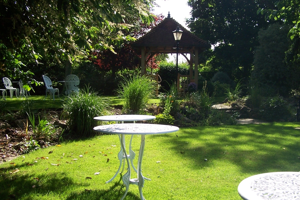 sunny garden with metal tables painted white.   flower beds in the middle, trees at the back with wooden pergola centre back.
